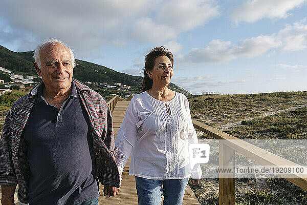 Happy senior couple holding hands walking on boardwalk under sky
