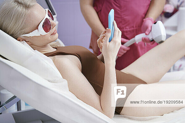 Woman using smart phone getting laser epilation treatment at clinic