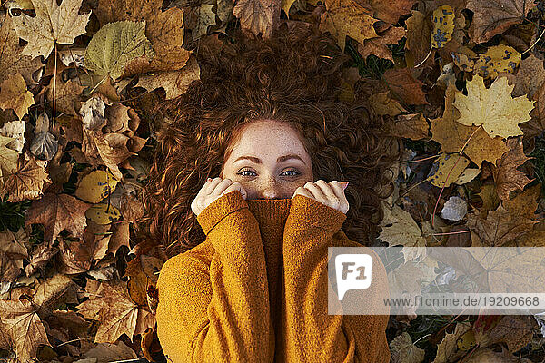 Redhead woman covering face with sweater lying on maple leaves at park