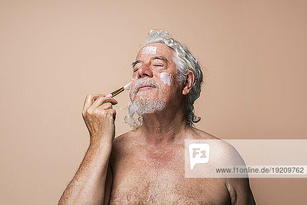 Senior man with eyes closed applying moisturizer using brush against beige background