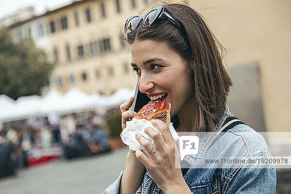 Smiling woman eating pizza and talking on smart phone