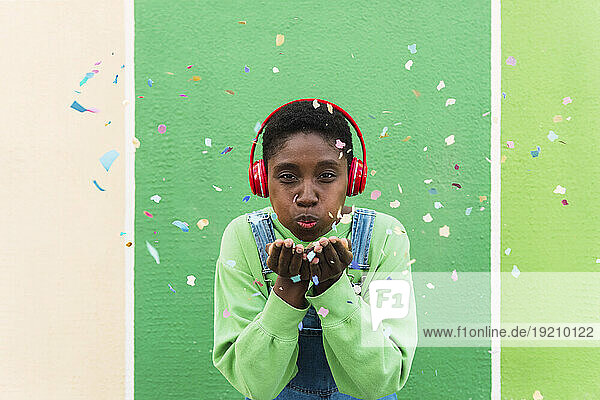 Young woman wearing wireless headphones blowing confetti in front of multi colored wall