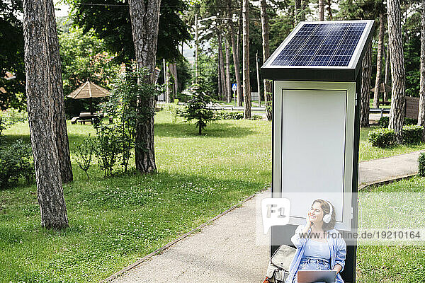 Smiling woman enjoying listening to music sitting near solar charging point at park