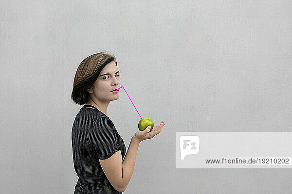 Teenage girl drinking from Granny Smith Apple with straw against gray background