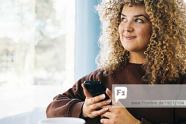 Thoughtful young woman with curly hair holding smart phone at cafe