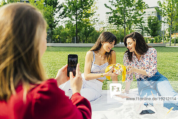 Woman photographing friends sitting on picnic blanket in park