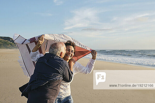 Senior man kissing happy woman holding scarf at beach