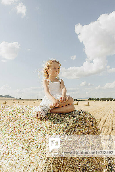 Blond girl sitting on bale of straw in field on sunny day