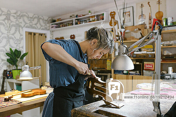 Luthier with hand tool carving on violin at desk in workshop