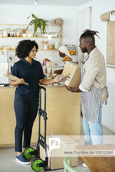 Smiling waiter holding package and delivery woman scanning QR code in cafe
