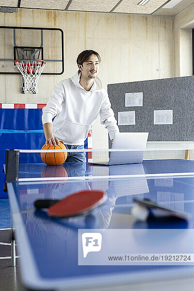 Smiling young businessman standing with basketball and laptop at tennis table
