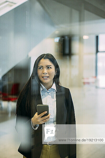 Businesswoman with smart phone seen through glass at workplace