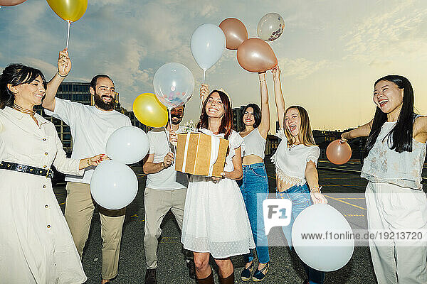 Cheerful friends celebrating woman's birthday with balloons and gift at sunset