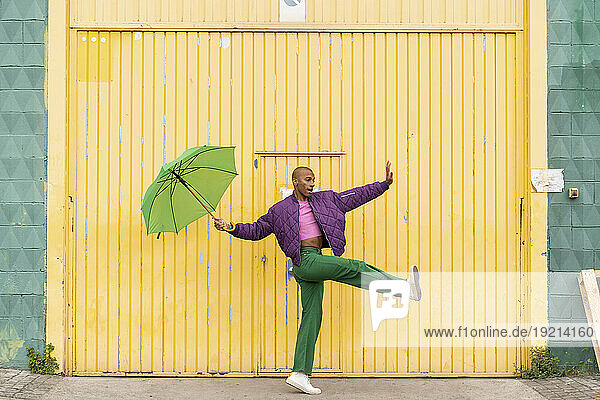 Non-binary person dancing with green umbrella in front of yellow shutter door