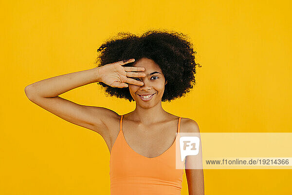 Smiling woman covering eye with hand against yellow background