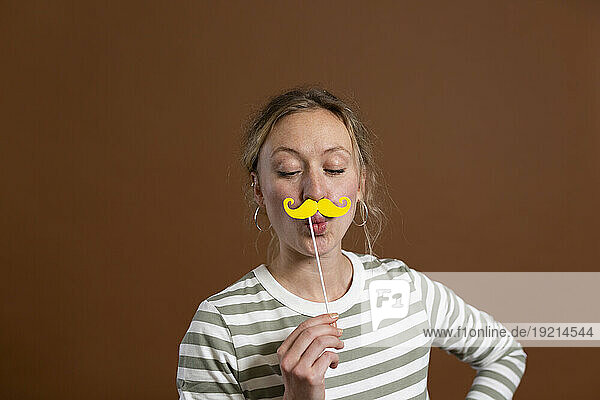 Woman puckering and holding mustache prop against brown background