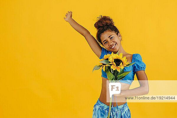 Happy woman with hand raised holding sunflower against yellow background
