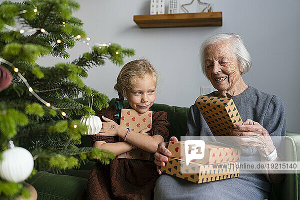 Happy granddaughter with grandmother holding gifts near Christmas tree at home
