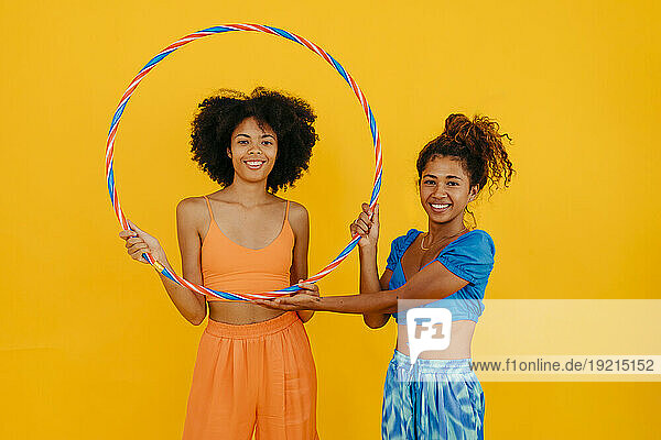Smiling women holding plastic hoop standing against yellow background
