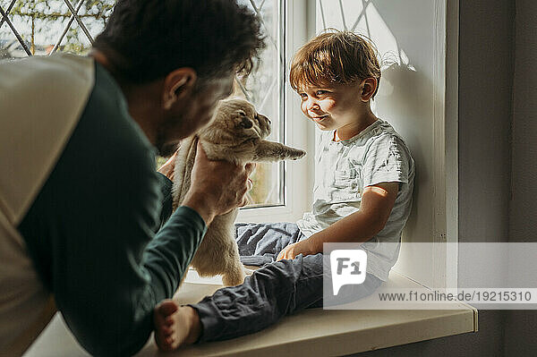 Cute boy looking at father with puppy in window at home