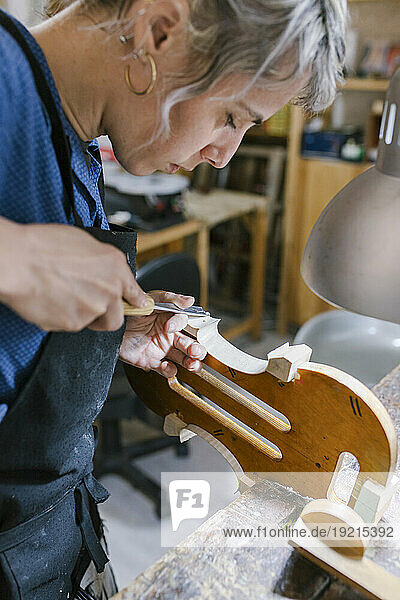 Luthier with chisel carving on violin at desk in workshop