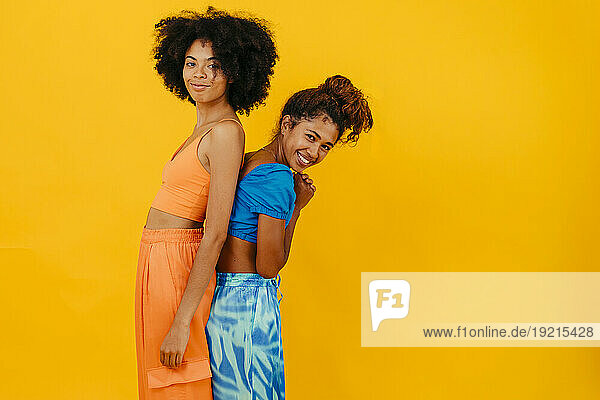 Smiling young friends standing back to back against yellow background