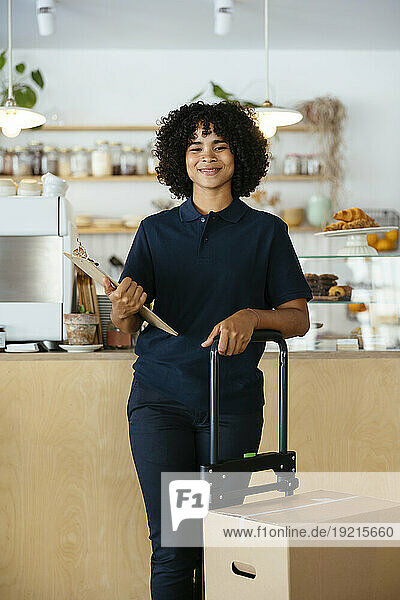 Smiling delivery woman holding clipboard and standing near package in cafe