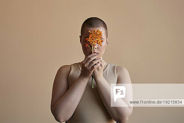 Woman covering face with orange flower against beige background