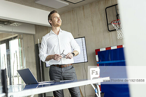 Happy businessman standing with laptop near tennis table in office