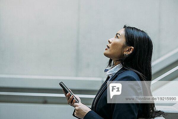 Businesswoman with smart phone looking up at workplace