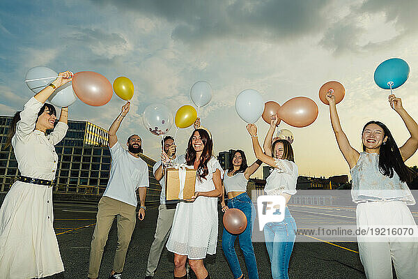 Happy friends celebrating woman's birthday with balloons and gift under cloudy sky at sunset