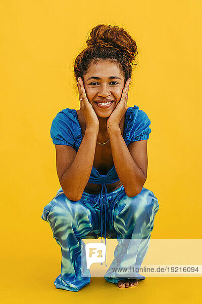 Smiling woman touching face crouching against yellow background
