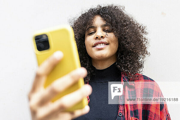 Smiling woman taking selfie with smart phone in front of wall