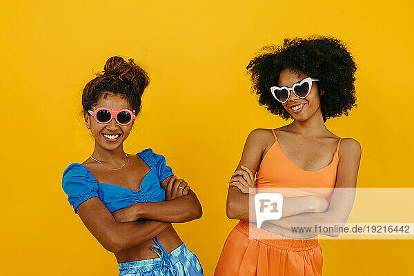 Women wearing sunglasses standing with arms crossed against yellow background