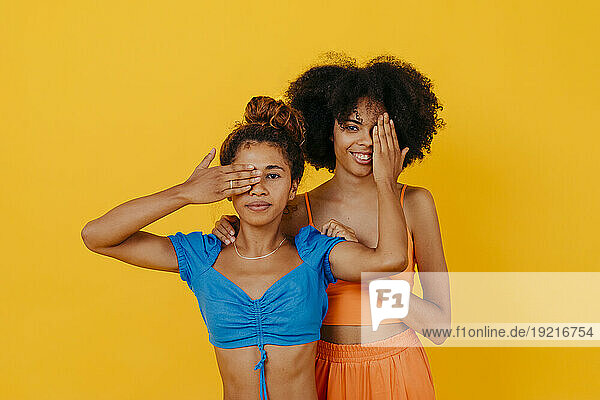 Woman covering eye of herself and friend against yellow background