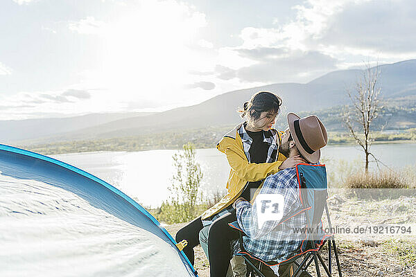 Girlfriend sitting on boyfriend's lap by tent near lake