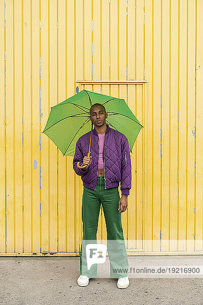 Non-binary person standing with green umbrella in front of yellow shutter door