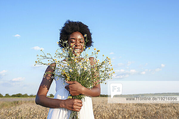 Smiling woman with eyes closed holding bunch of daises in field under sky
