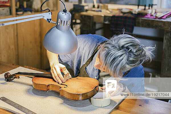 Instrument maker arranging bridge part on violin at desk in workshop