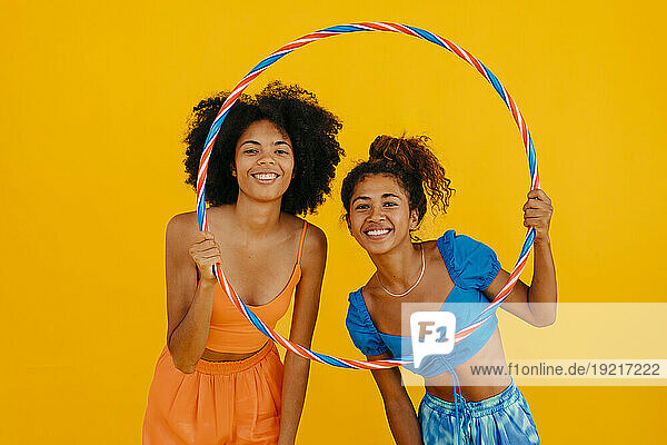 Happy young friends looking through plastic hoop against yellow background
