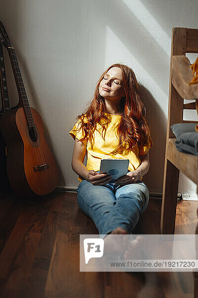Smiling redhead woman sitting with eyes closed and tablet PC on floor