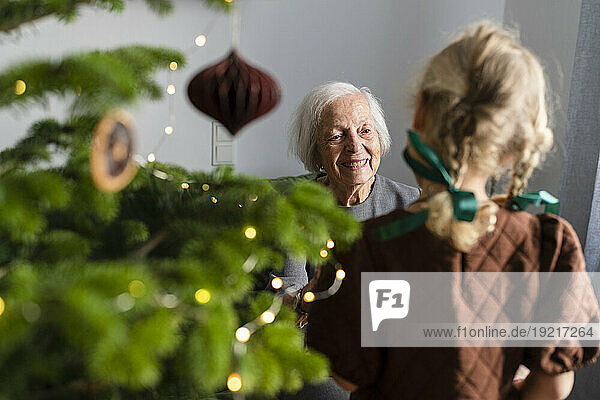 Smiling senior woman with granddaughter standing near Christmas tree at home