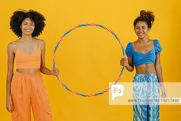 Smiling multiracial friends holding plastic hoop standing against yellow background