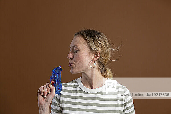 Blond woman blowing toy gun against brown background in studio