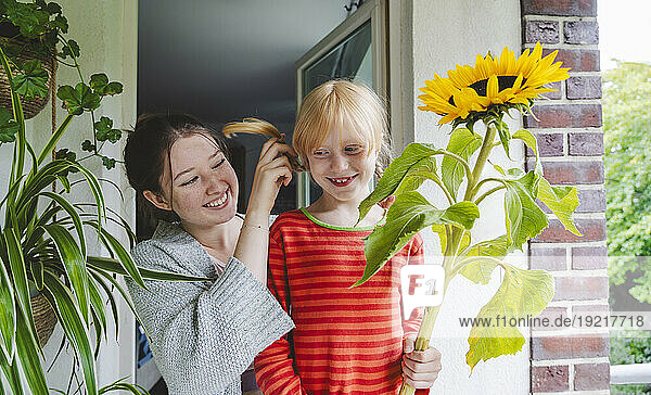 Smiling girl with big sunflower standing by sister on balcony