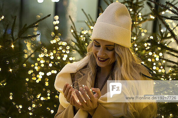 Smiling woman holding string lights near Christmas trees