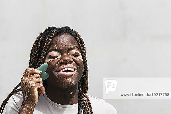 Happy young woman using gua sha in front of white wall