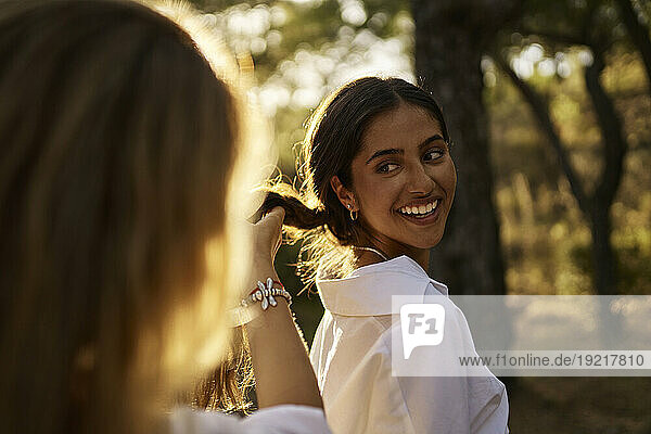 Happy teenage girl looking back at mother braiding her hair in forest