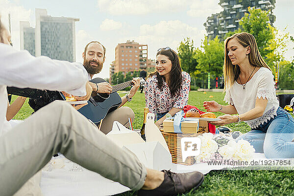 Smiling man playing guitar with friends enjoying picnic in park