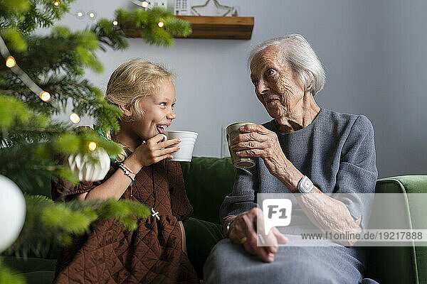 Happy girl drinking hot chocolate with grandmother and spending leisure time near Christmas tree at home